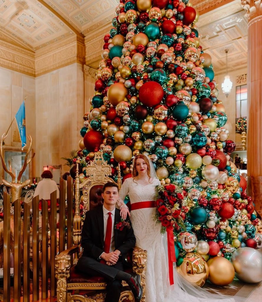 A couple wears red accessories and stands in front of a Christmas tree