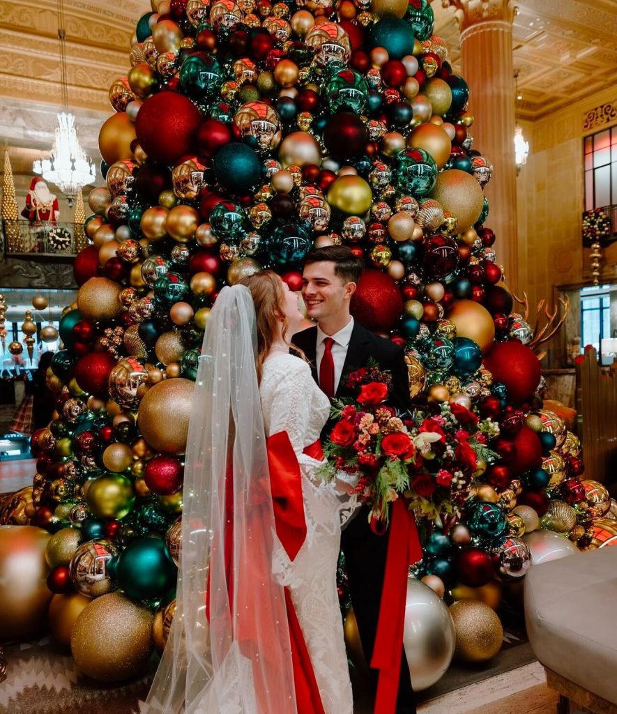 A couple wears red accessories and stands in front of a Christmas tree