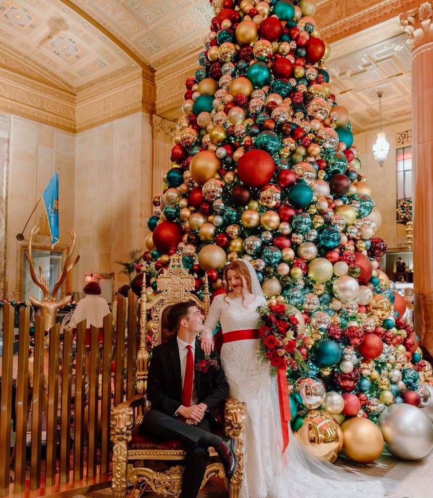 A couple wears red accessories and stands in front of a Christmas tree